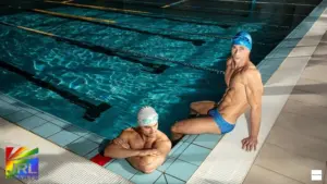 Male models wearing MODUS VIVENDI X Heart swimwear posing poolside in a modern indoor swimming pool setting